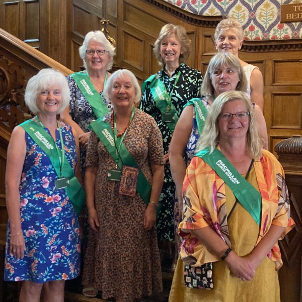 A group of women are standing on a staircase in a building. They are all wearing dresses and green Macmillan sashes.