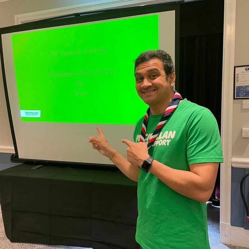 Kiwi is wearing a Macmillan branded top and jeans. He has short dark hair. He is standing and pointing towards a projector. The projector shows a presentation called 'My Cancer Journey: A Copper with Cancer by Kiwi'. 