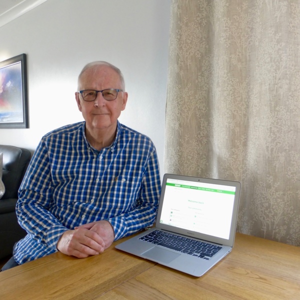 Michael is sitting down and leaning on a wood table. Michael is wearing a blue chequered shirt and glasses. On the table is a laptop that is open and shows the homepage for Macmillan's Online Community.