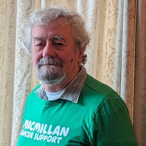 John has short hair and is seen from the waist up. He is wearing a green Macmillan branded top. He has a bear and moustache.