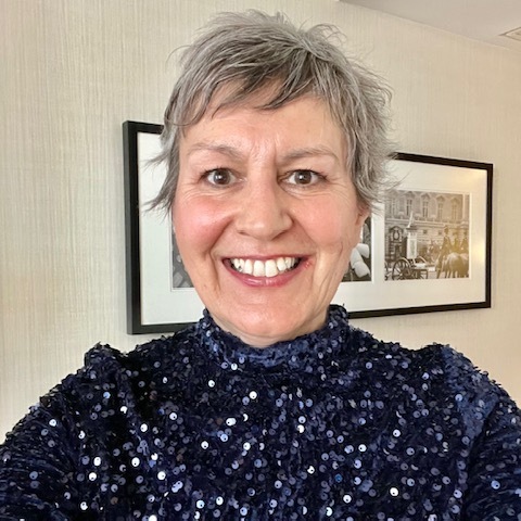 Allyson has short hair and is wearing a bright blue and sparkly top. She is smiling. Behind her is a wall with a large picture frame on it.