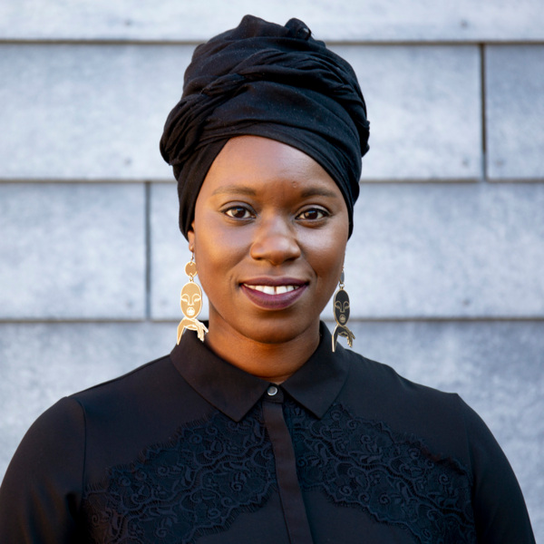 Sabrina is wearing a black top. She has a black hair wrap on and is wearing gold earrings. She appears to be outside.