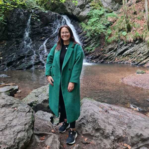 Jules is wearing a dark green jacket, a dark top and trousers, and dark blue trainers. She has long brown hair.  She is standing outside on some rocks near a waterfall.