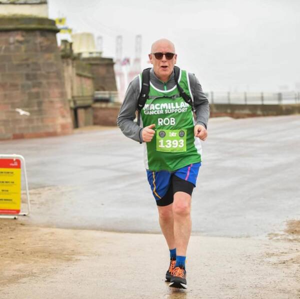 Rob is running outside. He is wearing black sunglasses, a green Macmillan branded technical vest, shorts and trainers. He is running on a cement path.