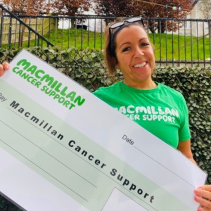 Ali is holding a large Macmillan branded cheque. She has her brown hair tied back. She is wearing a green Macmillan branded t-shirt and has a pair of glasses on the top of her head. Ali appears to be outside.
