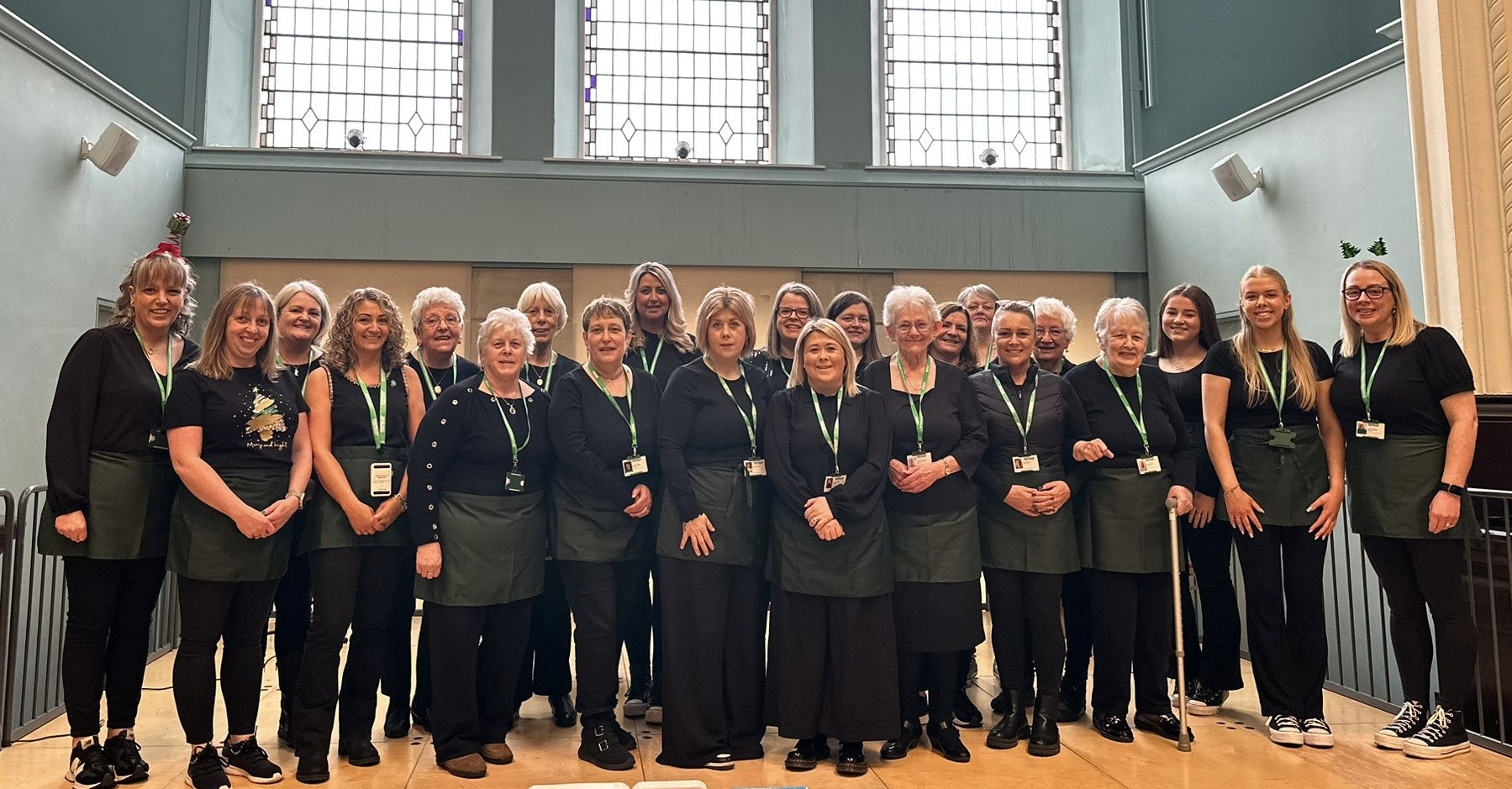 A large group of people in dark clothing are standing together. Some of them are wearing Macmillan branded lanyards. They appear to be indoors. 