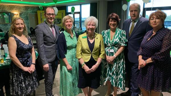 A group of 7 individuals are standing together in a row. They are indoors and it has been decorated with Macmillan branded decorations.