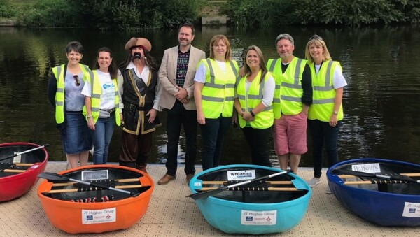 The Coracle Committee are standing in a row together outside. They are near a large body of water.