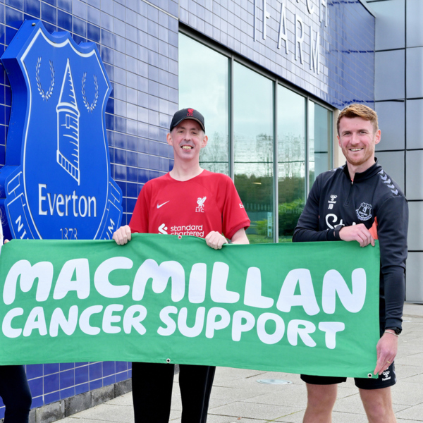 Jack it standing with 2 other people in a row. He is on the far right. He is holding a large Macmillan branded sign with the 2 other people. They are outside the Everton stadium. 