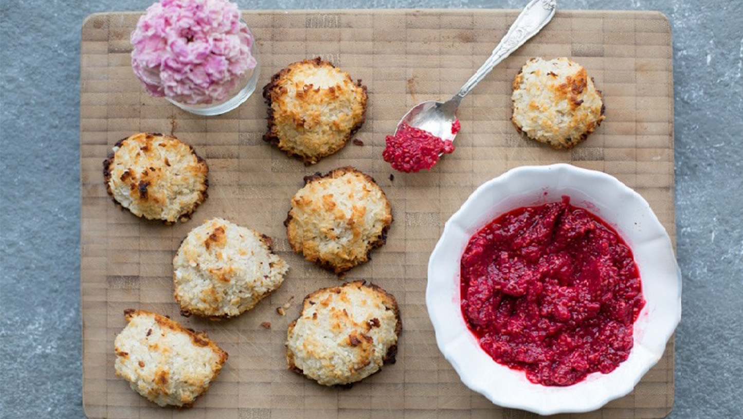 Seven golden-brown coconut macaroons with toasted edges rest on a wooden board, paired with a bowl of vibrant raspberry jam and a spoonful ready to serve.