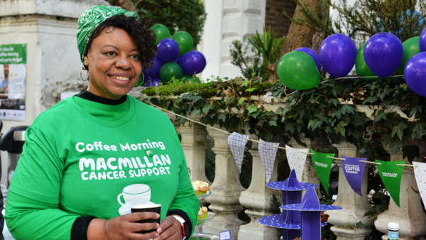 Debbie is wearing a green Macmillan branded Coffee morning t-shirt. She has hair hair in a green bandana too. She is standing outside holding a paper coffee cup. Behind her is a table decorated for a Coffee Morning event.