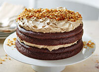 A decadent three-layer chocolate cake with rich, creamy salted caramel frosting between each layer and on top, crowned with crunchy peanut brittle pieces. Presented on a white cake stand. 