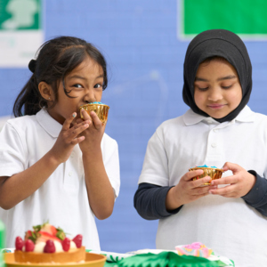 Two young people in school uniforms are holding cupcakes. The person on the left is biting into the cupcake. They appear to be in a school.