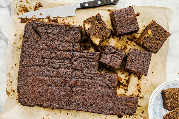 A rich, dark brown pear and ginger cake with a cracked surface sits on parchment paper, its moist texture revealed by square slices showing chunks of pear nestled in spiced ginger batter.