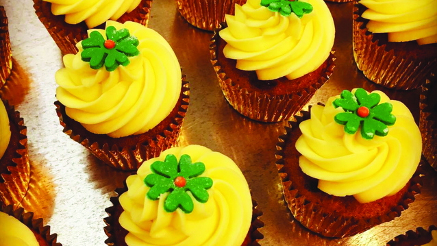 Rows of carrot cupcakes in metallic gold wrappers, each topped with a generous swirl of creamy yellow frosting and a delicate green fondant flower with a red center. 