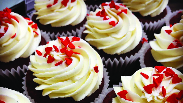 Red velvet cupcakes topped with generous swirls of creamy white frosting and decorated with red heart-shaped sprinkles. The rich red sponge peeks through beneath the frosting. 