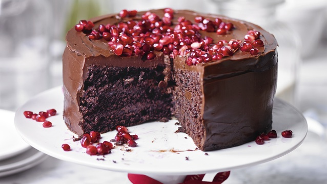 A rich chocolate cake with glossy chocolate frosting, topped generously with vibrant red pomegranate seeds. Displayed on a white cake stand with a large slice cut out, the cake reveals its moist, dense interior.