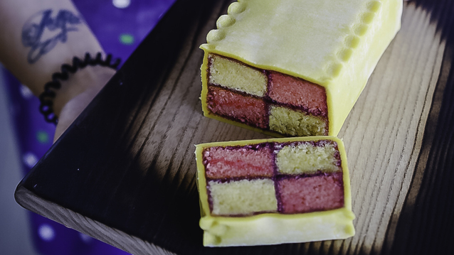 A Battenberg cake with a checkered pattern of pink and yellow sponge squares, bound with apricot jam and wrapped in smooth marzipan with a scalloped top edge. Presented on a wooden tray with one slice cut to reveal its vibrant interior.