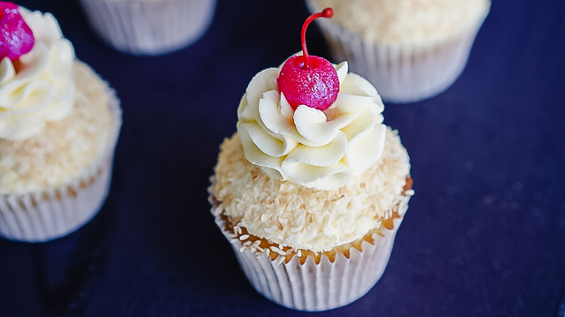 A golden-brown pineapple upside-down cupcake topped with creamy white frosting, a sprinkle of shredded coconut, and a bright red maraschino cherry with its stem.