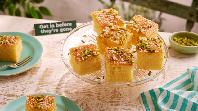 Six golden brown squares of moist revani cake on a glass plate and topped with shredded coconut and finely chopped pistachios.
