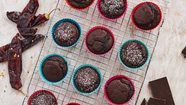 Many muffins are on top of a metal baking cooling rack. The cupcakes are in blue or red cupcake liners. Some of the cupcakes have a sprinkle of white powder on top. Next to the cooling rack are a few red chillies and a broken bar of chocolate. 