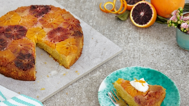 A polenta cake is on top of a square marble plate. A slice has been cut out of the cake. The slice is on a blue patterned plate. It has been topped with cream and an orange garnish. The plate also has a silver fork on it. 