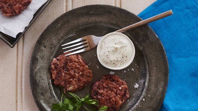 Two beetroot latkes are on a metallic plate. Next to them is a garnish, a pot with a white cream, and a silver fork. The plate is on top of a dark blue cotton napkin and a wooden table. There is also a baking tray with two latkes next to the plate. 
