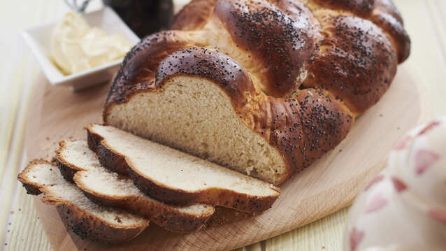 A loaf of challah bread is resting on top of a wooden serving board. Part of the loaf has been cut in slices. Also on the wooden board is an open jar with a spoon resting on top of it and a small plate with butter in it. 