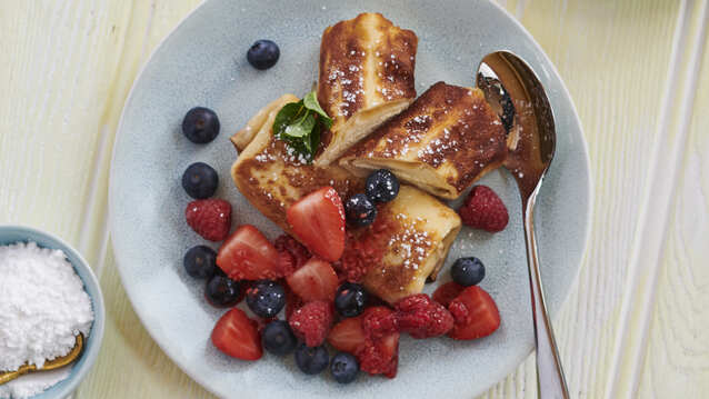 Two cheese blintzes are on a light blue plate. One of the blintzes has been sliced in half. They are topped with strawberries and blueberries, and a white dusting. There is also a silver spoon on the plate. 