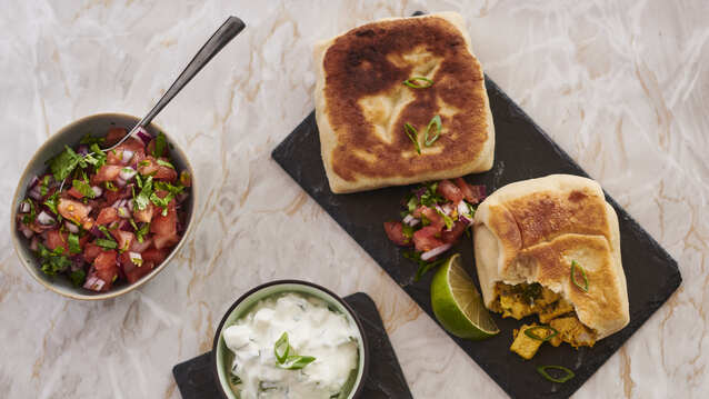 Two dark rectangular slate plates are on top of a light marble table top. On the plates are rotis. On one plate a roti has had a bite taken out of it. Next to the plates is a small bowl with chopped vegetables that has a spoon in it. One plate also has a pot of a white dip in it. 