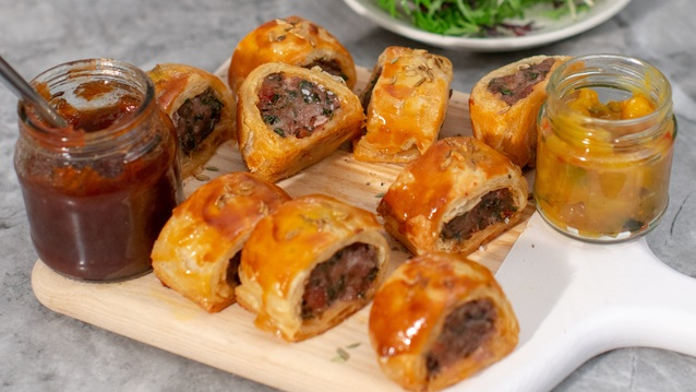 Several chorizo sausage rolls are on a wood and plastic board. They are jars of condiments also on the board. Behind them is a plate with different leaf vegetables. 