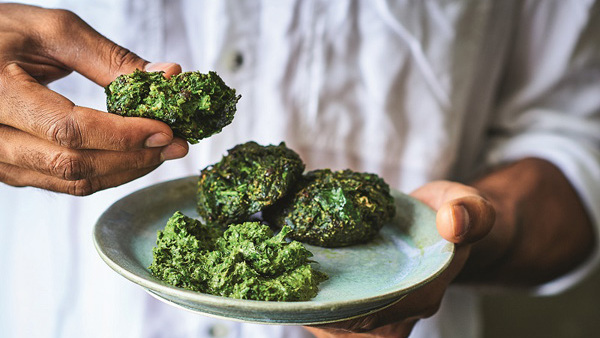 A person in a white shirt is holding a pakroa in one hand and a plate of pakoras in the other. The person is only seen from the shoulders down. The pakroas are green and the plate is a light blue. 