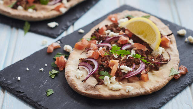 Two lahmacuns are each placed on rectangular slate plates. They are topped with different vegetables and cooked minced meat. 