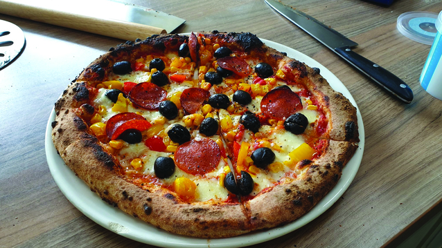 A pizza topped with pepperoni, black olives, cheese and pineapple is on a white plate. The pizza has been cut into pizza slices. Next to the pizza is a knife. 