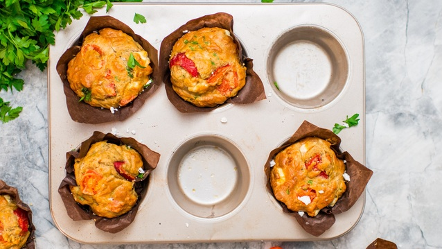 A view of muffins from above. Some muffins are placed in a muffin tin, and some are on the surface where the tin is placed. The muffins are wrapped in brown parchment paper. Next to the muffins and tray is a bundle of parsley and some cut up tomatoes. 