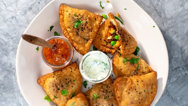 Several vegetable samosas are on a white plate. There is also a jar of a white sauce and a chutney on the plate. The chutney jar has a utensil sticking out of it. 