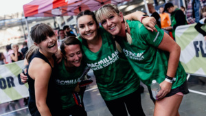 Four people are standing in a row, slightly bent over, smiling and with their arms wrapped around each other's shoulders. Some of the group are wearing Macmillan branded vest tops. They appear to be outside at an event.