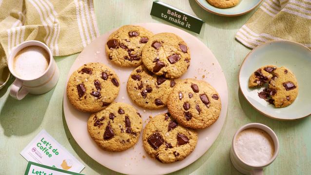 A plate of golden-brown chocolate chip cookies with gooey melted chunks sits on a light green wooden table. Two frothy coffees in white and beige mugs, partially eaten cookies on side plates, and green-striped napkins create a cozy, inviting scene.