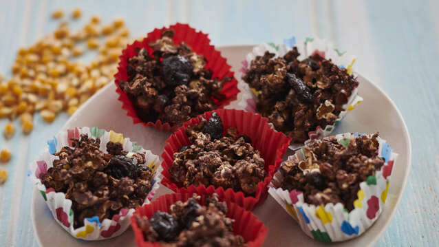 Six rich chocolate and dried fruit clusters nestled in vibrant cupcake liners—red, floral, and polka-dotted—sit on a plate. Packed with oats, chocolate, and chewy dried fruits, their textured surface looks indulgent and satisfying.