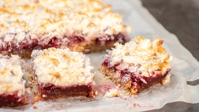 A freshly baked dessert bar with a light crust, vibrant red berry filling, and toasted coconut flakes sits on parchment paper. One piece is cut out, revealing its moist, crumbly layers—an irresistibly sweet and fruity treat.