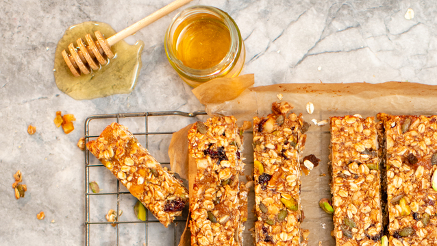 A batch of freshly baked granola bars packed with oats, nuts, seeds, and dried fruits rests on a cooling rack. Rectangular pieces are cut and ready to serve, while a jar of golden honey with a dripping dipper adds a glossy, indulgent touch.