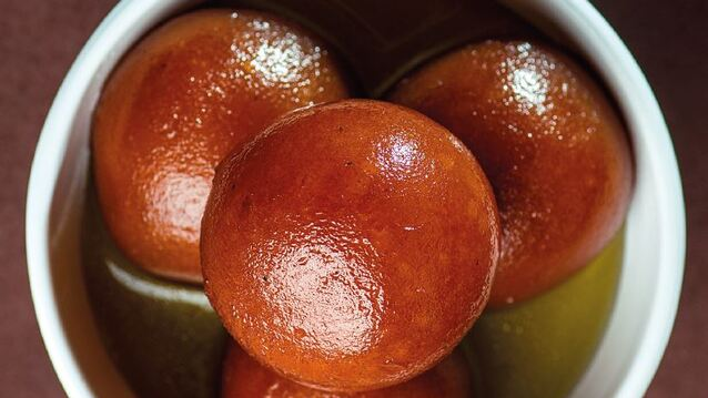 Four golden-brown spherical gulab jamuns glistening in sugar syrup. Their glossy, syrup-soaked surface and rich color make them look irresistibly moist, sweet, and indulgent.