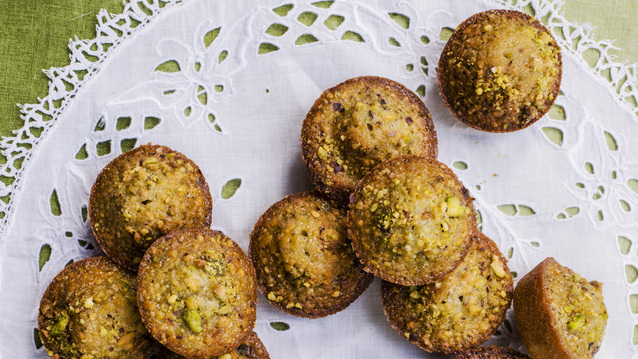 A batch of freshly baked pistachio mini muffins with golden-brown crusts and crunchy green pistachio pieces, arranged on a white lace doily. Set against a green tablecloth, the close-up view makes these nutty treats look irresistibly warm and inviting.