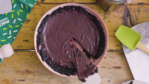 A decadent dark chocolate tart with a glossy surface and Oreo cookie crust sits on a rustic wooden table. A slice is being lifted out, revealing its creamy, rich filling.
