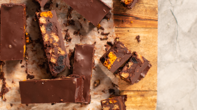 Slices of rich, glossy chocolate-covered treats rest on a wooden surface, revealing chunky bits of biscuit or cookie inside. The smooth chocolate coating glistens under the light, with scattered crumbs making them look irresistibly indulgent.