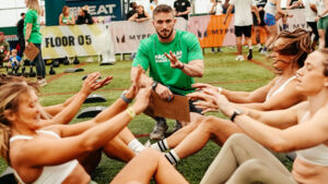 People in groups of 2 are sitting across from each other and doing sit ups. They have their legs locked together. A person in a green Macmillan branded top is squatting down near the group. He is holding up 2 fingers and has a clip board.