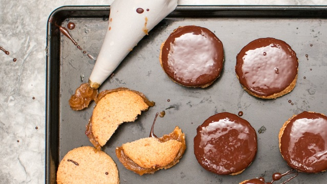 A baking tray filled with round, golden cookies—some topped with glossy chocolate. A piping bag oozes rich caramel in the corner, white crumbled cookie pieces reveal a soft, crumbly texture.