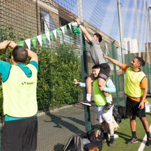 A group of footballers are on a football pitch putting up bunting on a metal fence. One footballer in a grey top is sitting on top of another's shoulders.