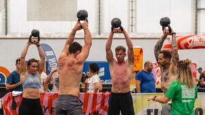 A group of people are competing in an exercise at a Battle Cancer event. They are standing in a circle and holding a dumbbell with both arms over their heads. A person in a green top is holding a clipboard and observing the group.