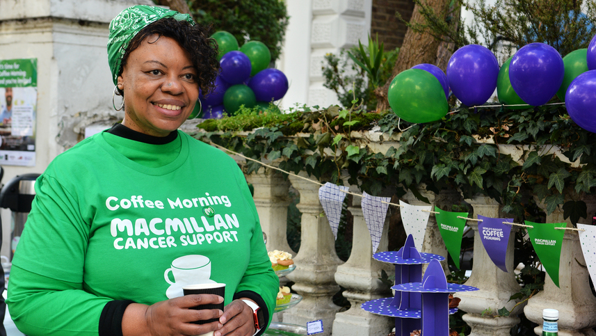 Debbie is wearing a green Macmillan branded Coffee morning t-shirt. She has hair hair in a green bandana too. She is standing outside holding a paper coffee cup. Behind her is a table decorated for a Coffee Morning event.